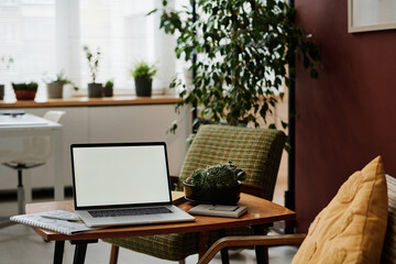 Laptop with white screen mockup placed on table in modern office with greenery and vintage furniture