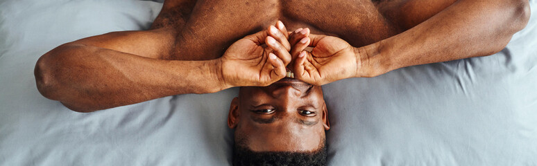 joyful african american guy covering mouth and smiling at camera waking up on bed, top view, banner