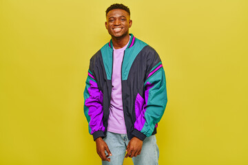 cheerful african american man in colorful windbreaker jacket looking at camera on yellow background