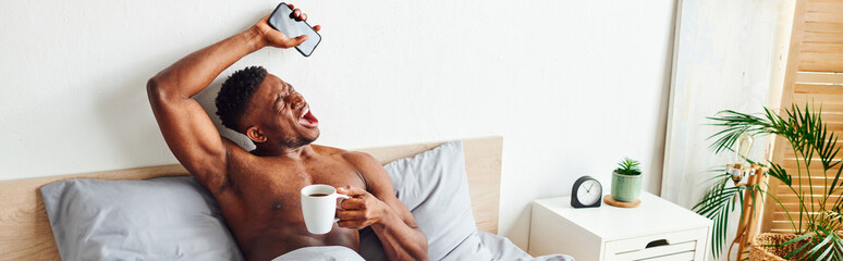 muscular african american man with coffee cup and smartphone yawning and stretching on bed, banner