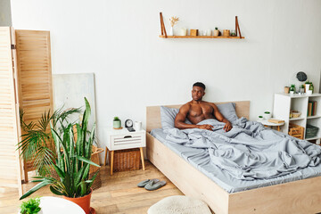 shirtless muscular african american man sitting on bed in spacious bedroom with green potted plants