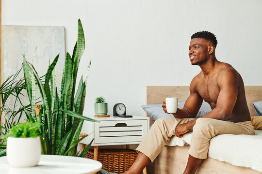 smiling shirtless african american man with coffee cup sitting near green potted plants in bedroom