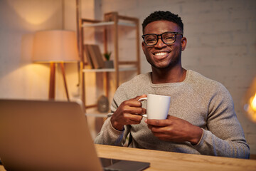 cheerful african american freelancer with coffee cup sitting near laptop at night in home office