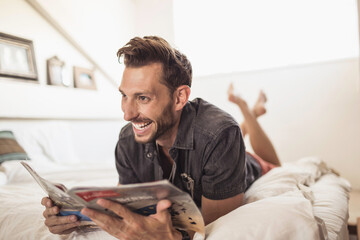 Man at home lying in his bed reading a magazine and smiling.