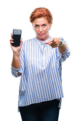 Atrractive senior caucasian redhead woman showing smartphone screen over isolated background pointing with finger to the camera and to you, hand sign, positive and confident gesture from the front