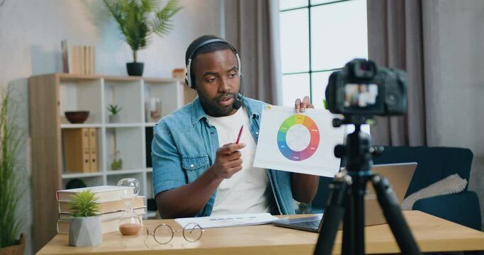 Attractive confident African American man motivated in headset recording video on professional camera at home and explain colorful diagram for his audience