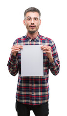 Young hipster adult man holding blank paper sheet scared in shock with a surprise face, afraid and...