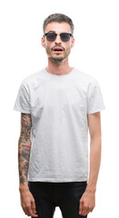 Young tattooed adult man wearing sunglasses scared in shock with a surprise face, afraid and...