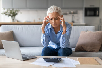 Frustrated senior woman calculating finances touching head in despair indoor