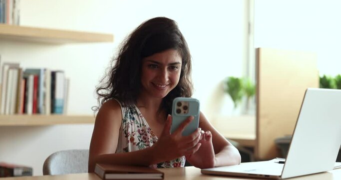 Smiling Indian woman hold smartphone at workplace, sit at desk with laptop, take break chat with friend, answer to client use messenger application, make call by business, enjoy online communication