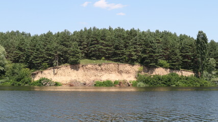 Fototapeta na wymiar A cliff made of clay near a wide river. A coniferous forest grows above the cliff.