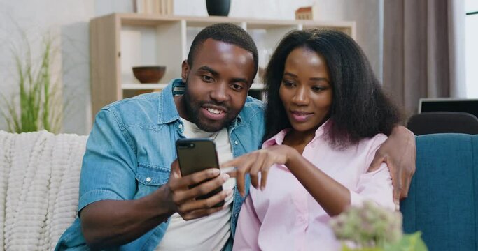 Happy loving African American man and woman sitting together on comfortable sofa at home and browsing mobile apps,family relationship concept