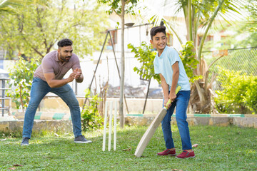 Cheerful indian father encouraging son from behind while playing cricket game at park - concept of...
