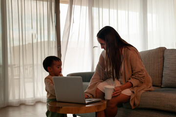 Mom talking to baby while working at home on laptop, coffee cup in hand.