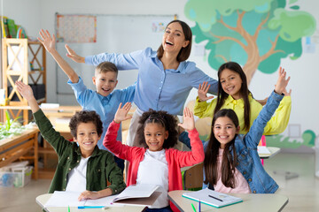 Excited diverse schoolchildren raising arms and smiling, sitting in classroom with their happy female teacher