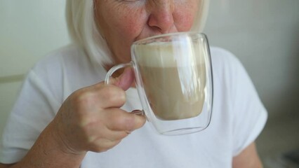 Close up of Caucasian 60s woman sitting by the window drinking coffee latte at kitchen. Beautiful woman enjoy drinking coffee at cafe. 