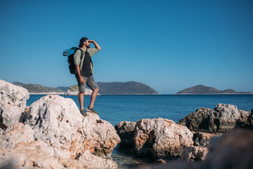 A male tourist with a large backpack and hiking gear on a rocky seashore.