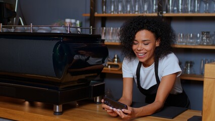 Dark-skinned beautiful woman plays games on her phone while working. The barista is distracted from...