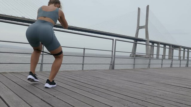 Young woman in sportswear training glutes muscles with resistance band with a river and skyline in the background, focused and determined.