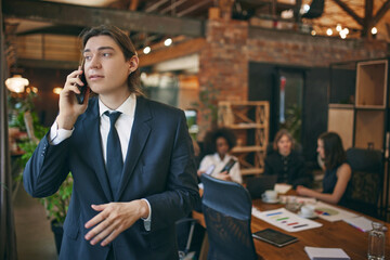 Portrait of confident man, student in official clothes talking on phone while preparing for exam in public library. Selective focus. Concept of study, co-working space, teamwork, collaboration. Ad
