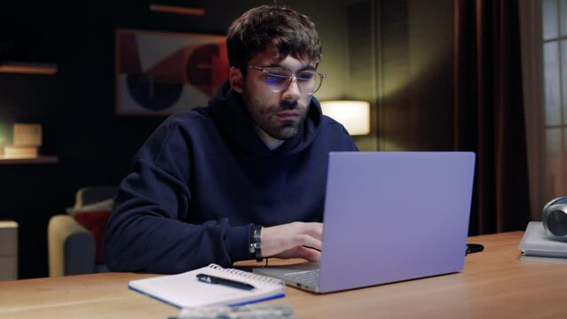 Man sitting at table and using laptop. Bearded male looking at laptop screen. Man satisfied with result of work. Male holding pen and revising finished project. Man spending evening at home.