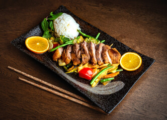 Asian food. Peking duck, rice and vegetables on a black ceramic plate, on the background of a wooden table