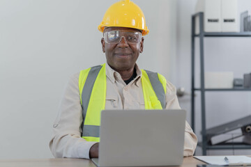 American construction site manager standing with folded arms wearing safety vest and helmet, thinking in the office. Used laptop for checking job satisfaction.