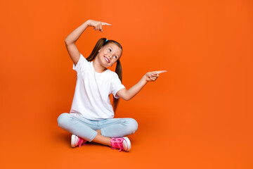 Full body photo of cute positive girl sit floor look indicaate fingers empty space ad isolated on orange color background