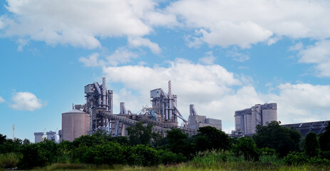 Obraz premium View of cement industrial factory with trees and cloudy sky background, Thailand. Environment conservation in business concept.