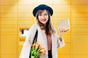 Portrait of smiling fashion woman with box near modern postal automatic mail terminal with self...