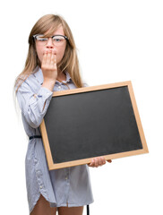 Young blonde child holding blackboard cover mouth with hand shocked with shame for mistake, expression of fear, scared in silence, secret concept