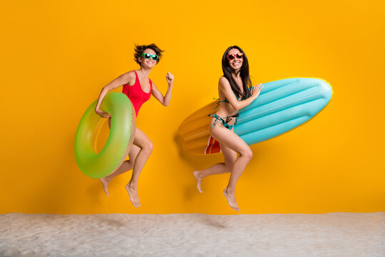 Full length photo of excited funky ladies wear bikini jumping high running swimming buoys isolated yellow color background