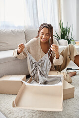 joyous attractive african american woman unpacking her new chic silver dress and smiling joyfully