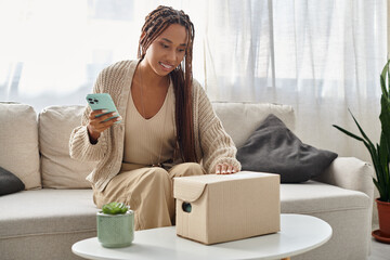 joyous good looking african american woman holding smartphone and looking at cardboard parcel