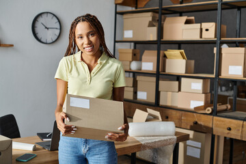 appealing hard working african american female seller holding box and smiling at camera, delivery