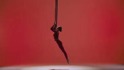 Zelfklevend Fotobehang Female gymnast isolated on red studio background. Girl aerial dancer in bodysuit on acrobatic trapeze with straps perform element. © kinomaster