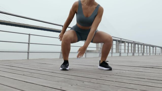 A determined 25-year-old woman exercises outdoors on a cloudy day, mastering Floor Touch Squat Jacks for strength and agility