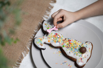 Hand of child putting pink bow on ear of baked Easter bunny gingerbread cookie with multi colored sugar sprinkles and chocolate bunny tail on served table. Playing with food, spending time with family