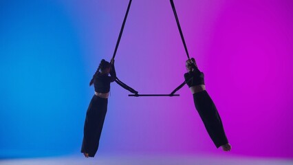 Female gymnastic duet isolated on neon studio background. Girls aerial dancers performers spin on acrobatic trapeze with straps.