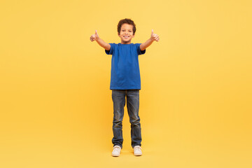 Fototapeta na wymiar Cheerful boy giving thumbs up in blue shirt on yellow background