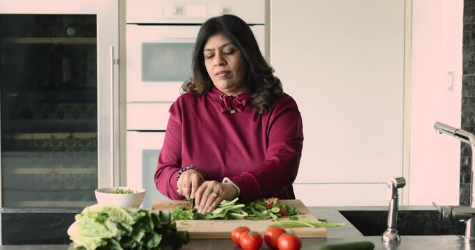 Smiling middle-aged Indian woman housewife cutting lettuce for dietary dish for family dinner or lunch, cooking alone in domestic kitchen, keep healthy lifestyle. Culinary, homemade food preparation