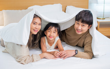 Three people of Asian family, father, mother, little daughter laying down under or covered by blanket on bed in bedroom at cozy home, smiling with happiness. Kid, Housing, Healthcare Concept.