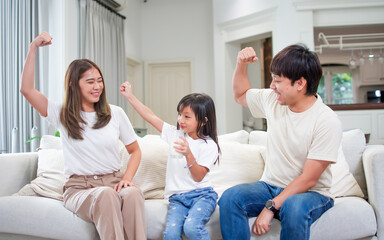 Three people of Asian family, father, mother, little daughter serving, drinking milk in living room at cozy home, smiling with happiness, posing by showing muscles for good health. Kid Concept.