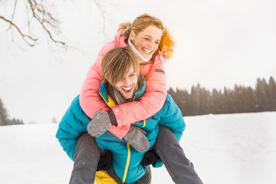 Man carries his wife piggy back across a field in the snow in winter. Wolfratshausen, Bavaria, Germany