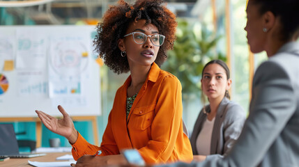 Female professionals are engaged in a discussion, with one of them gesturing expressively, likely explaining or presenting a concept with a whiteboard full of graphs and data in the background.