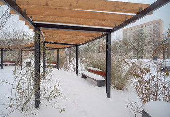 Mały osiedlowy park w zimowej aurze . Ozdobna pergola, nad spacerową alejką z ławkami , oraz...