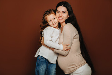 Studio portrait of young happy family pregnant woman and cute little daughter, girl gently hugging mother expecting child