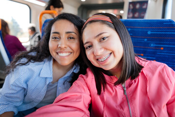 Two young cheerful latin student women traveling on public train transportation in the city and taking selfie with phone