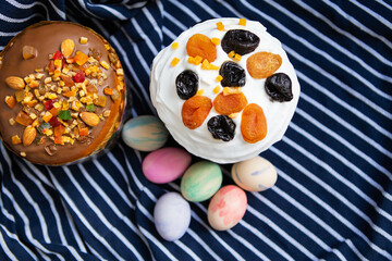 Delicious Easter cakes with colorful eggs on striped fabric. Two cakes, one with white frosting, the other with chocolate frosting and mixed nuts. Multi-colored painted eggs with different patterns.