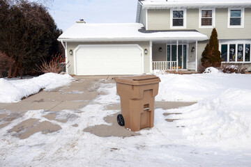 Plastic trash bin on the driveway of a suburban home in the snow, view from the street